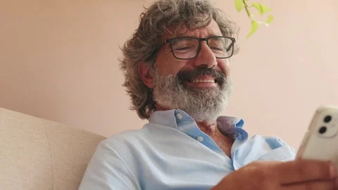 Close up, smiling elderly man sitting on sofa in living-room with phone in his Stock Footage 255962982