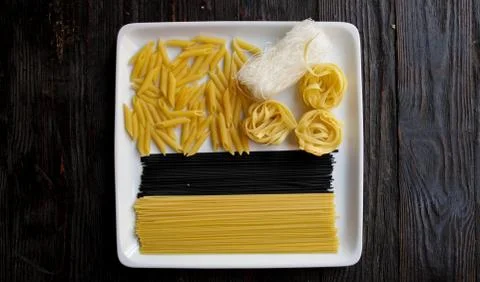 Close up of square white plate with various types of dry pasta.Dark wooden ba Stock Photos