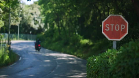 Close up of a stop sign beside a peaceful green road, with a motorcycle passing Stock Footage 339758474