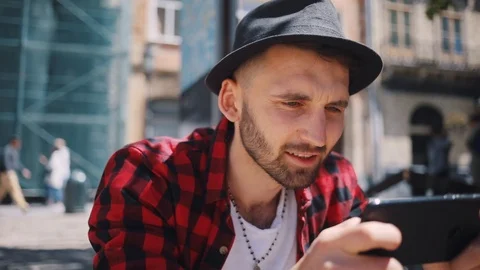 Close up stylish handsome young man in the hat uses phone play games sitting in Stock Footage 92072019
