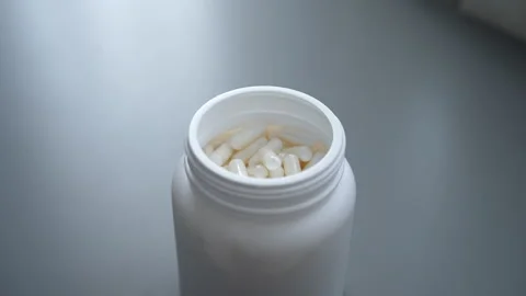 Close-up tracking shot of white pills filling white pill jar placing on gray Stock Footage 297953785