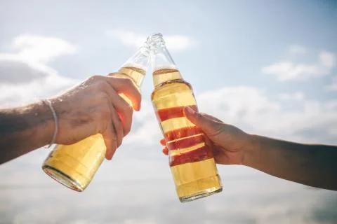 Close-up two bottles with beer in hands in the sunshine against the sky Stock Photos