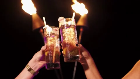 Close up of two drinks clinking glasses, cheers in front of lit tiki torches. Stock Footage 135238039