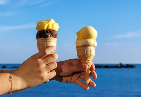 Close up of two hands holding ice cream cones against a blue sea and sky ba.. Stock Photos