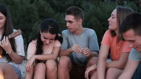 Close view of friends sitting around bonfire, playing ukulele on sandy beach Video stock 166819262