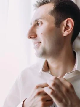 Close-up view of the groom buttoning his shirt and looking through window. Stock Photos
