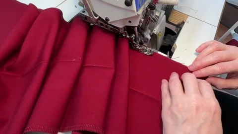 A close-up view of hands as they guide piece of red fabric through serger Stock Footage 296074571