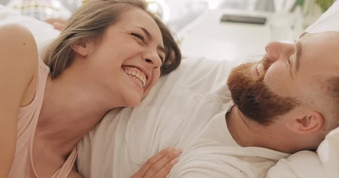 Close up view of happy couple lying on bed in early morning, talking andlaughing Stock Footage 127346256