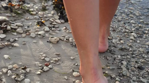 A close-up view of legs walking along beach with small pebbles, water gently Vidéo 282902656