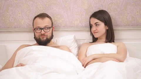 Close-up view of a loving couple in bed, a girl lifts a blanket and looks  스톡 동영상 102398716