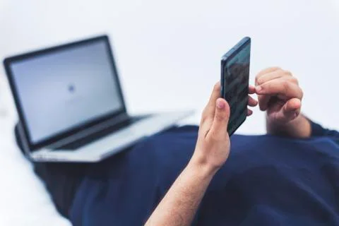 Close up view of a man using his mobile phone and a lap top while lying on be Stock Photos