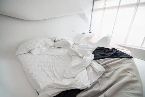 Close view of a rumpled white bed with a dark throw blanket in a curved, fu.. Stock Photos