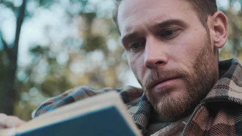 A close-up view of a serious man is reading a book sitting in the autumn park Vídeos de archivo 154042706