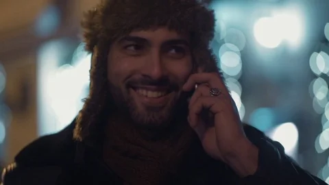 Close up view of smiling bearded man in a fur hat talking via phone. Night city Video stock 84652795