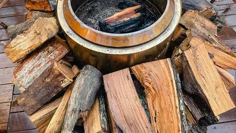 Close up of a warm metallic fire pit surrounded by chopped firewood and wet.. Stock Photos