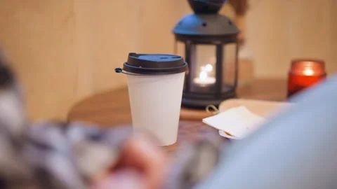 Close up white disposable coffee cup black lid wooden table lit lantern candle Stock Footage 347979284