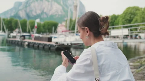 Close-up, woman sitting on pier using mobile phone on lake and mountains 스톡 동영상 290153221