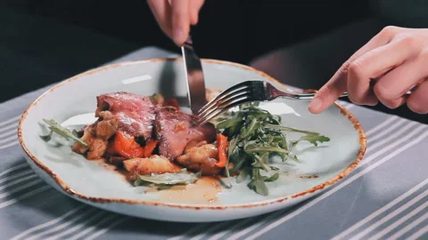 Close up woman's hands with knife and fork cutting meat in dish on table in a Stock Footage 88021434