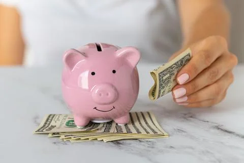 Close-up of a woman's hands put dollar paper money in a piggy bank, next to.. Stock Photos