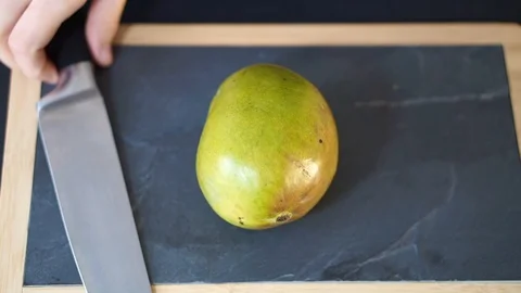 Close up, a woman's hands take a knife and cut the Mango exactly in half Stock Footage 106760136
