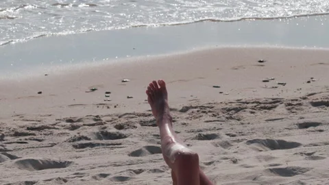 Close up of a woman's legs stretched on the beach as gentle waves roll in Stock Footage 320572790