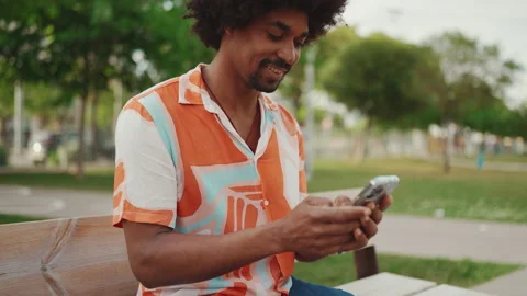Close-up of young African American man wearing shirt sitting on park bench using Stock Footage 227046341
