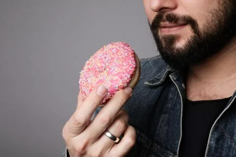 Close-up of young bearded man biting pink donut piece with pleasure. Fotos Stock