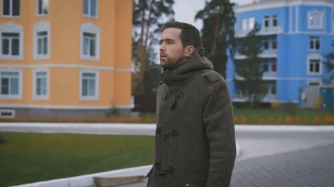 Close up of young bearded man in brown coat walking in the street near buildings Stock Footage 70463454