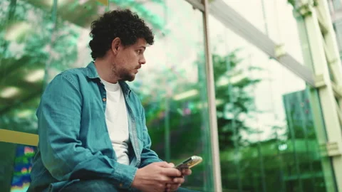 Close-up, young bearded man in denim shirt sits at bus stop waiting for bus and Stock Footage 226984021