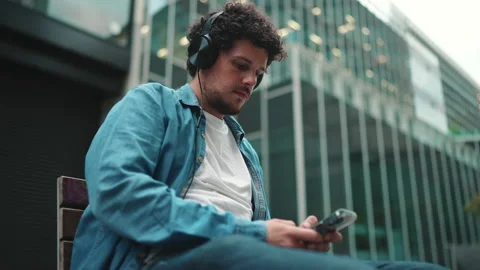 Close-up of young bearded man in denim shirt sitting in headphones on bench Stock Footage 226986130