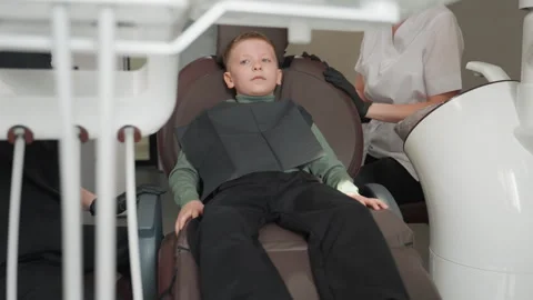 Close up of young boy lying in dental chair with black bib as dentist assistant Stock Footage 345834471