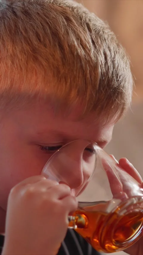 Close up of young boy steadily sipping from clear glass with amber liquid in Stock Footage 345728756