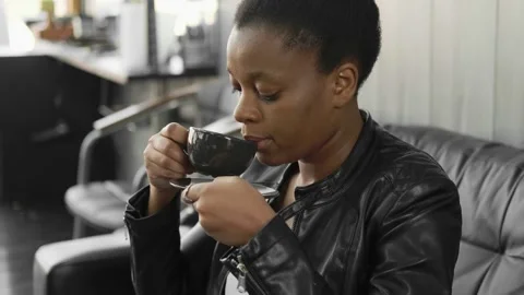 Close-up. Young calm black woman relaxing while sitting on a black sofa and drin Stock Footage