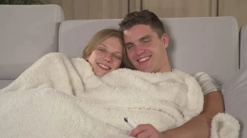 CLOSE UP: Young couple cuddling under blanket on a sofa and watching TV program Stock Footage 219374122