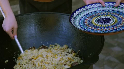 Close-up of young female cook serving Uzbek plov from large cauldron onto 스톡 동영상 346589056