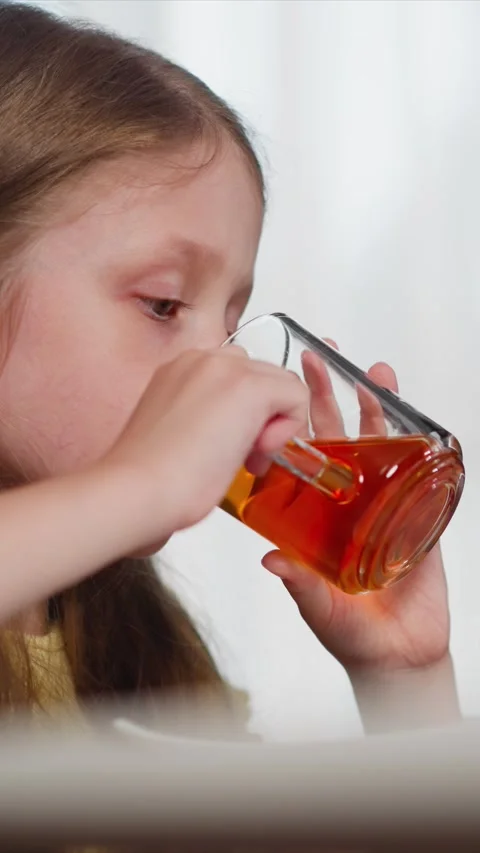 Close up of young girl drinking tea from clear glass with amber liquid in soft Stock Footage 344791100