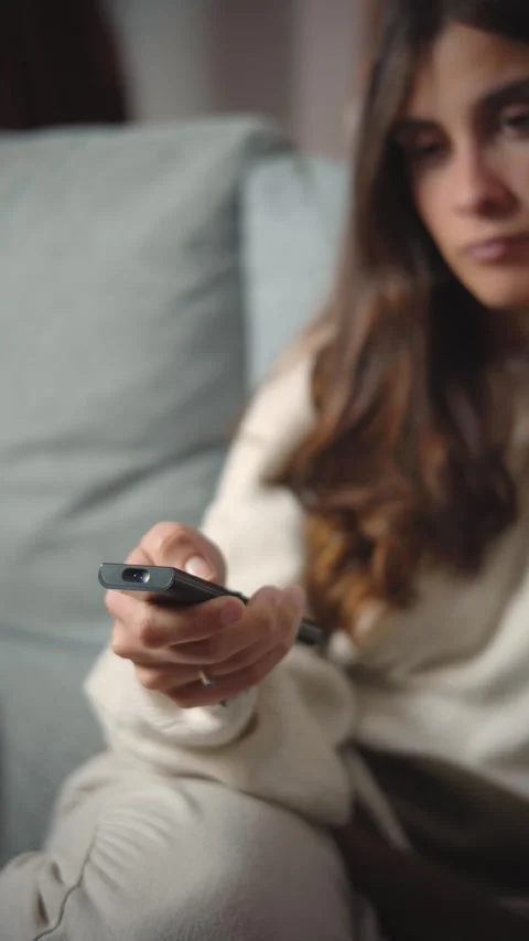 Close-up of a young Hispanic woman's hand using a TV remote while sitting on her Stock Footage 346162383