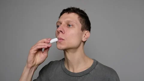 Close-up of a young man applying lip balm on dry lips against a gray background. Stock Footage 296883320