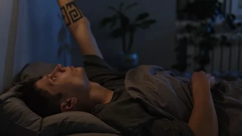 Close-up of a young man in bed, feeling sleepy after reading a book, turning off Stock Footage 296056676