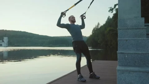 Close up young man exercising with trx suspension strap near lake crossfit Stock Footage 138650121