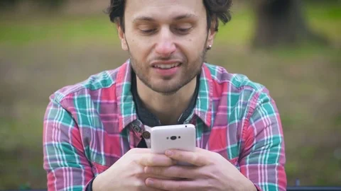 Close up on young man sitting on a bench at the park, chatting with his phone Stock Footage 74718942