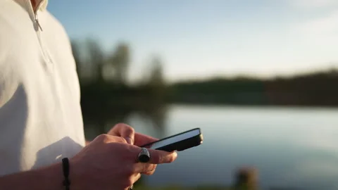 Close-Up of Young Man Using Smartphone by Lake at Sunset Stock Footage 339031402