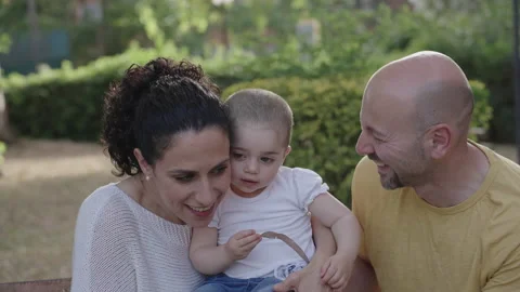 Close-up of young parents playing and kissing their little girl on a park bench Stock Footage 226678050
