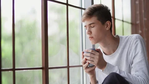 Close-up. A young sad guy sits by the window and drinks Stock Footage 154715875