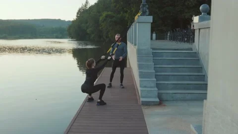 Close up young training squats with trx near lake at sunrise with trainer man Stock Footage 138651879