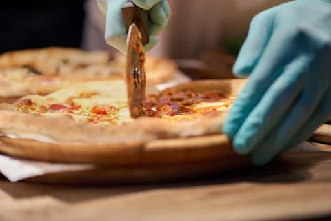 Closeup hand of chef baker in blue gloved hand, cutting pizza at kitchen Stock Photos
