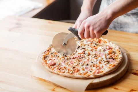 Closeup hand of chef baker in uniform blue apron cutting pizza at kitchen Stock Photos