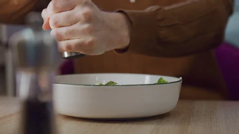 Closeup hand grinding pepper over fresh salad leaves in white bowl on wooden 動画素材 345731275