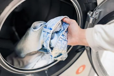 Closeup of a hand loading blue denim jeans into the drum of an open washing.. Stock Photos