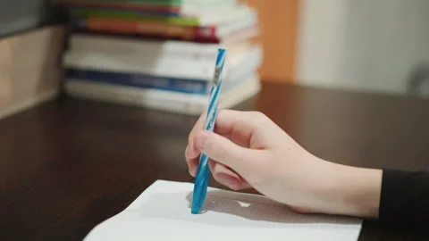 Closeup hand writing on notebook with blue pen, stacked books on desk in soft Stock Footage 332274273